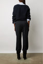 Person standing with their back to the camera, wearing a dark textured sweater, light shirt, black pantalon de tailleur, and black heeled boots, against a plain white background.