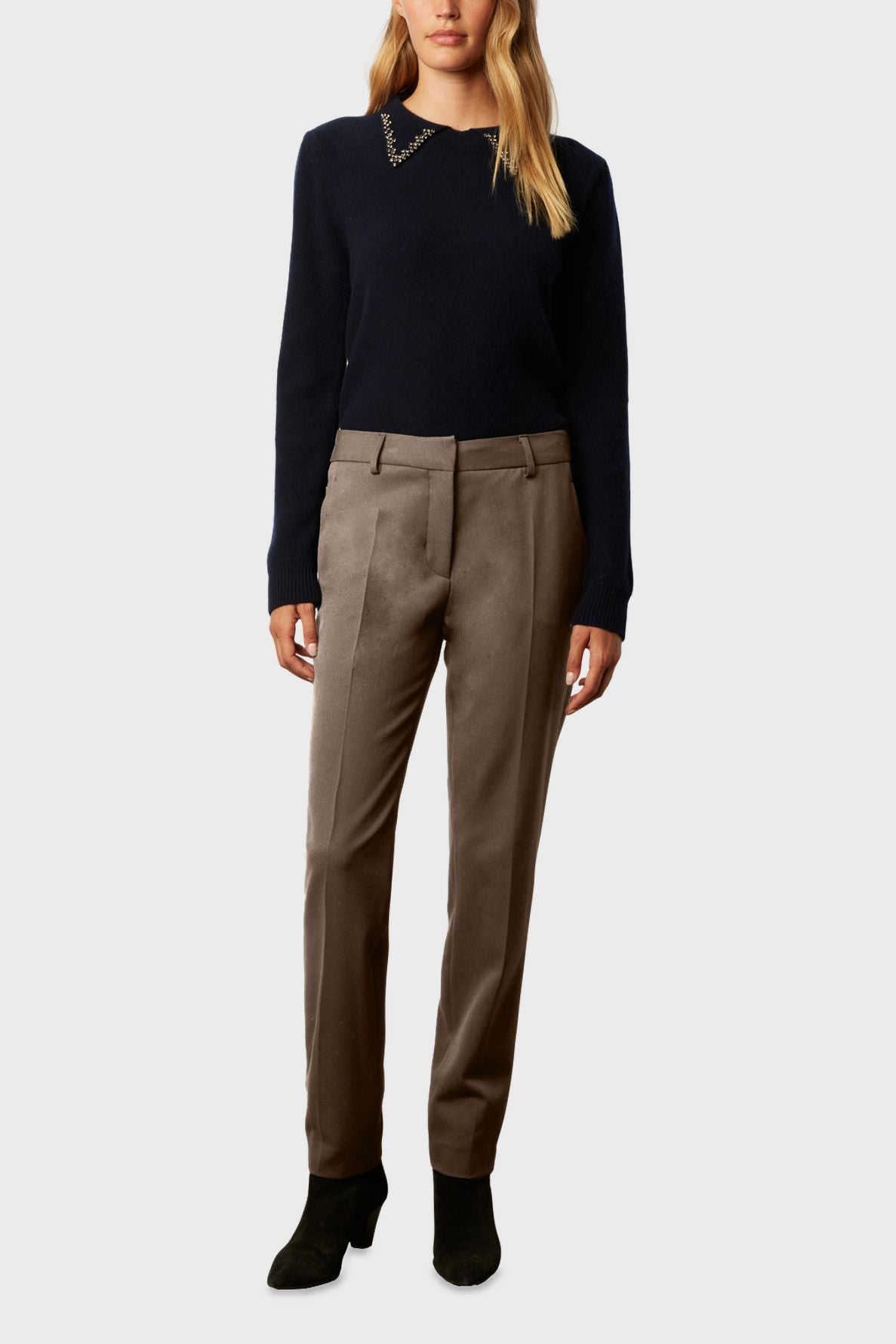 Woman wearing a navy sweater with chain detail on the collar, taupe dress pants, and black heeled ankle boots, standing against a plain light background. Her head is partially out of frame.