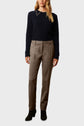 A woman wears a navy sweater with a small chain detail at the collar, taupe tailored pants, and black heeled boots. She stands against a plain light background, facing forward with her arms relaxed.