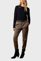 A woman stands with hands in pockets, wearing a navy sweater, taupe tailored pants with pleats, a black belt, and black ankle boots. She has long, wavy hair and a neutral expression.