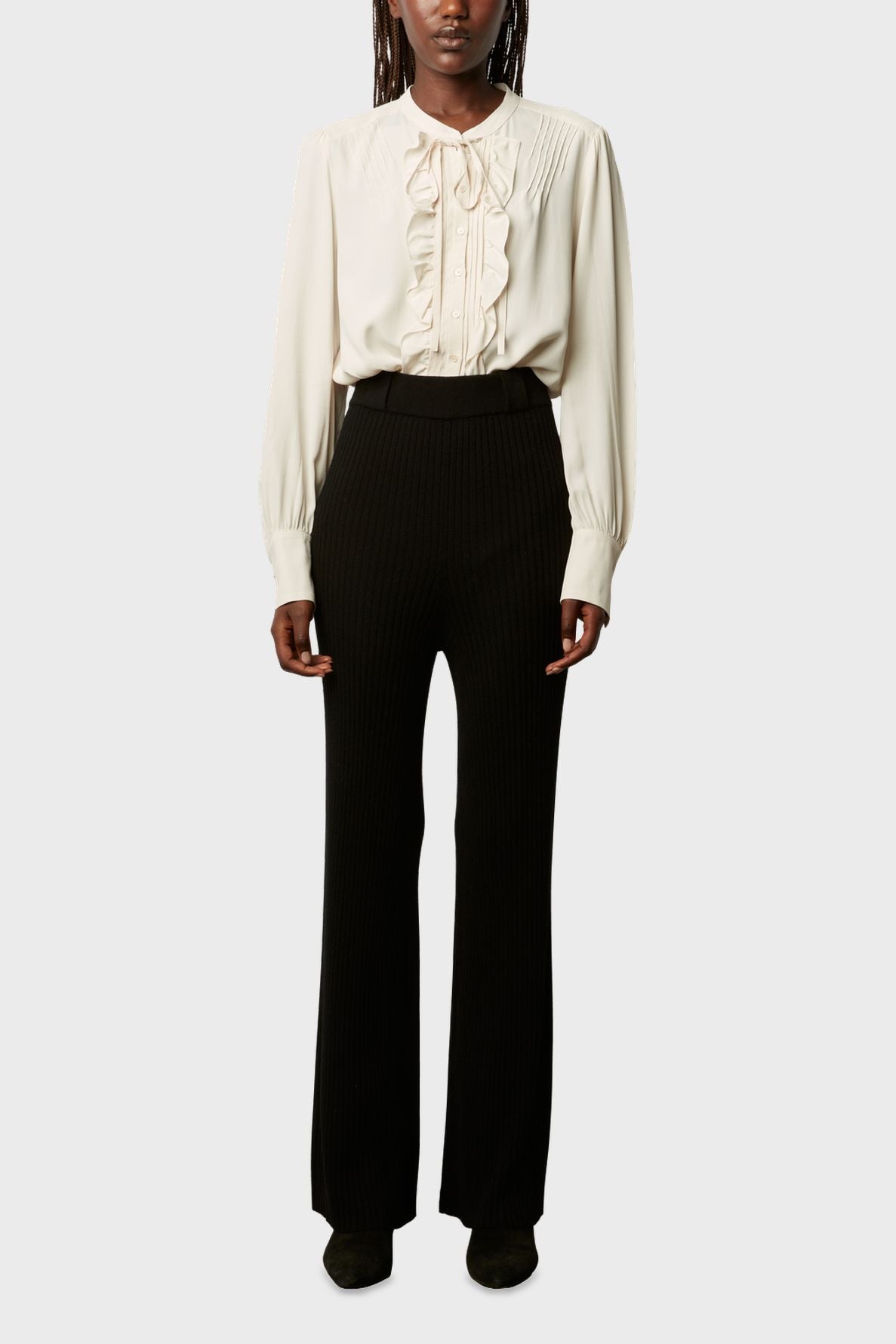 Person wearing a cream long-sleeve blouse with ruffles down the front, paired with a black pantalon large taille haute in laine mélangée, and black shoes, standing against a plain light background.