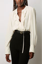 Woman wearing a cream ruffled blouse with long sleeves, tucked into a high-waisted pantalon large in black ribbed fabric, accessorized with a black belt featuring a gold buckle. Only torso and part of face are visible.