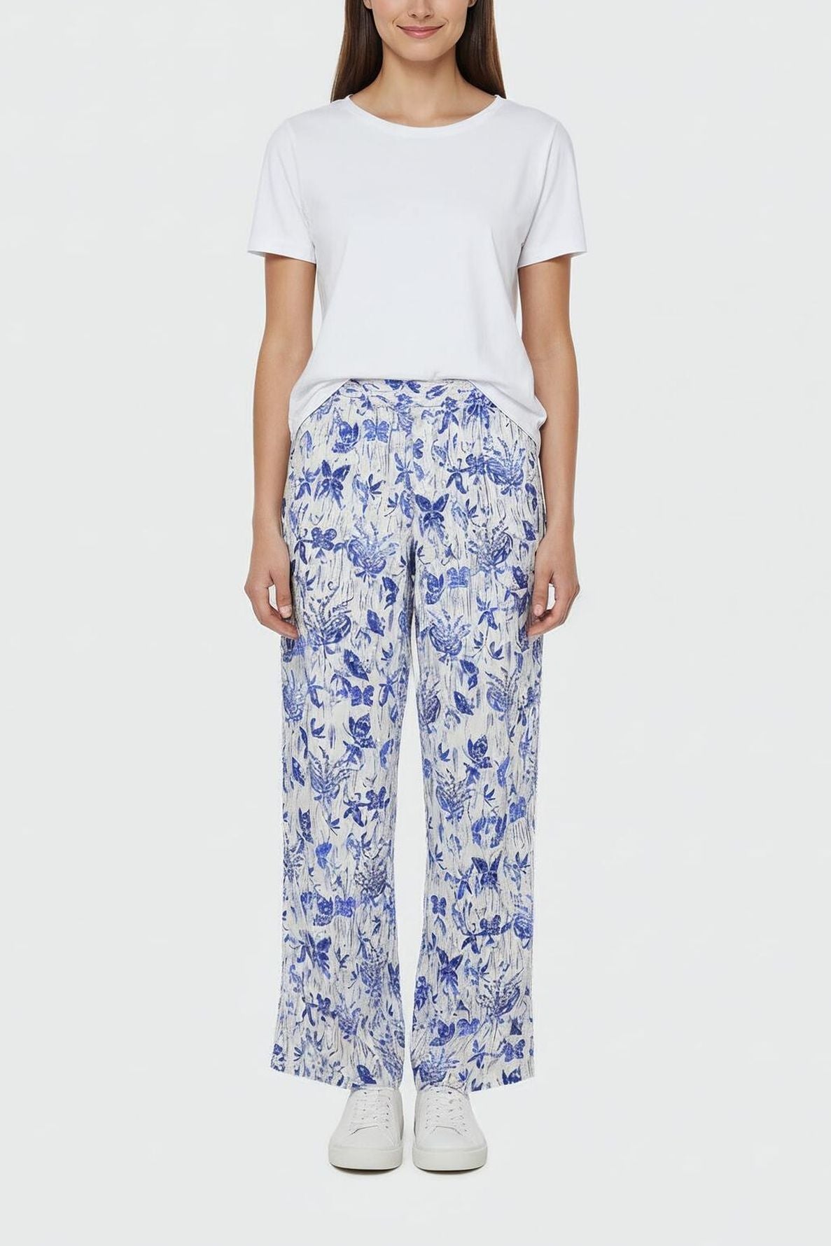 A woman stands facing forward wearing a plain white t-shirt, loose taille haute white pants with an imprimé fleuri bleu, and white sneakers, against a plain white background.