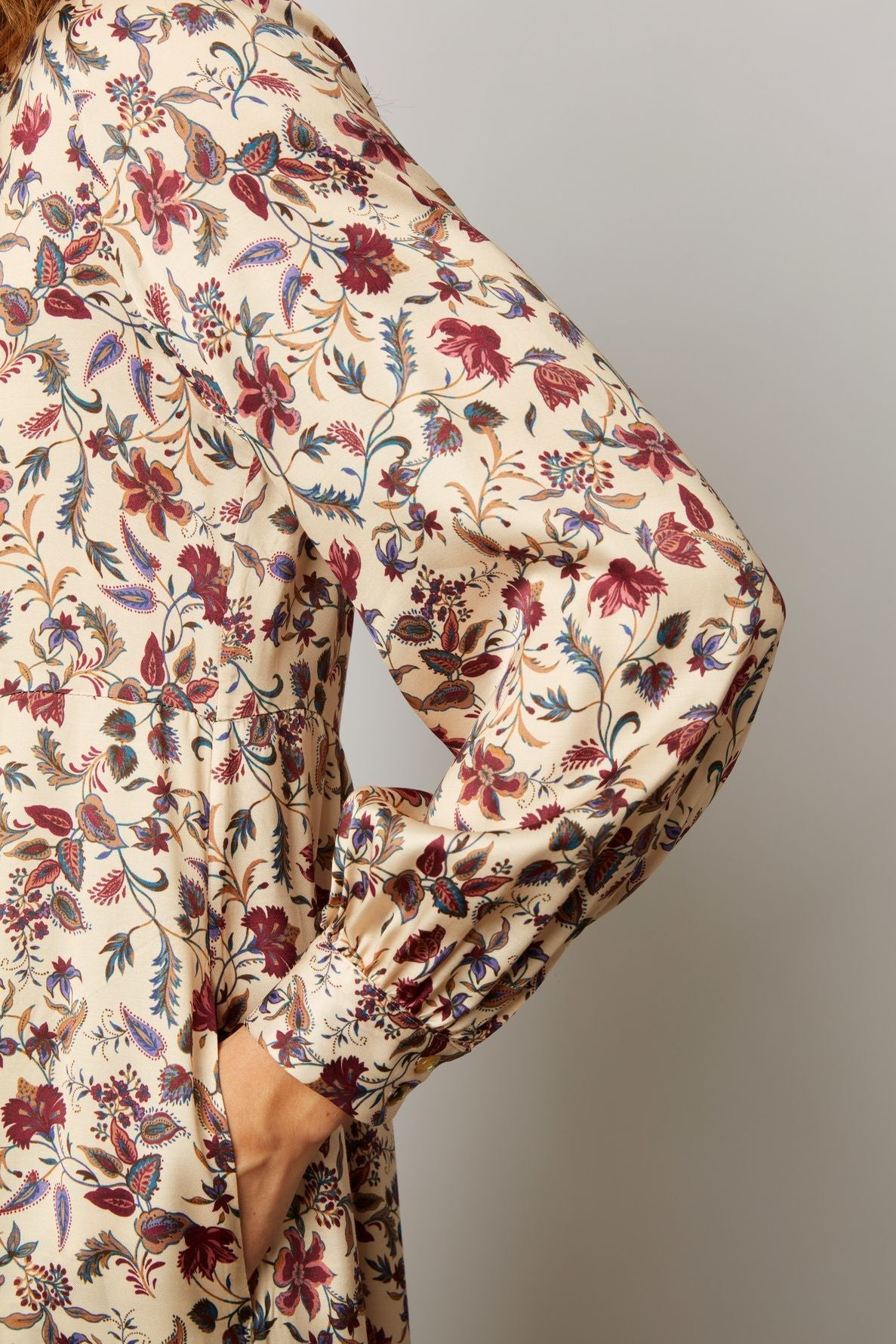 A person wearing a cream-colored, long-sleeved dress with a red and blue floral pattern poses with one hand on their hip against a plain background.