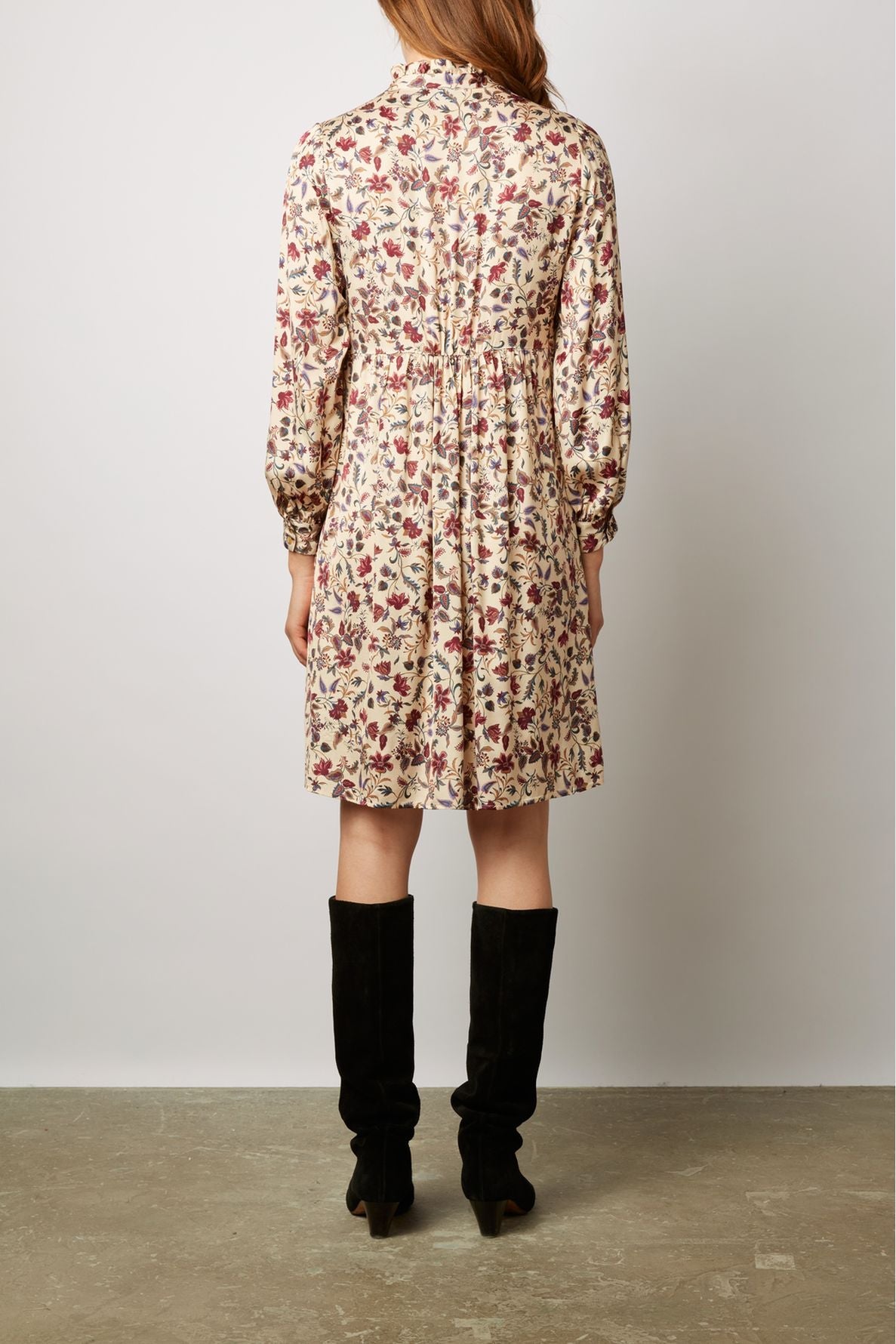 Woman wearing a cream-colored, floral-patterned long-sleeve dress and black knee-high boots, standing with her back to the camera against a plain background.