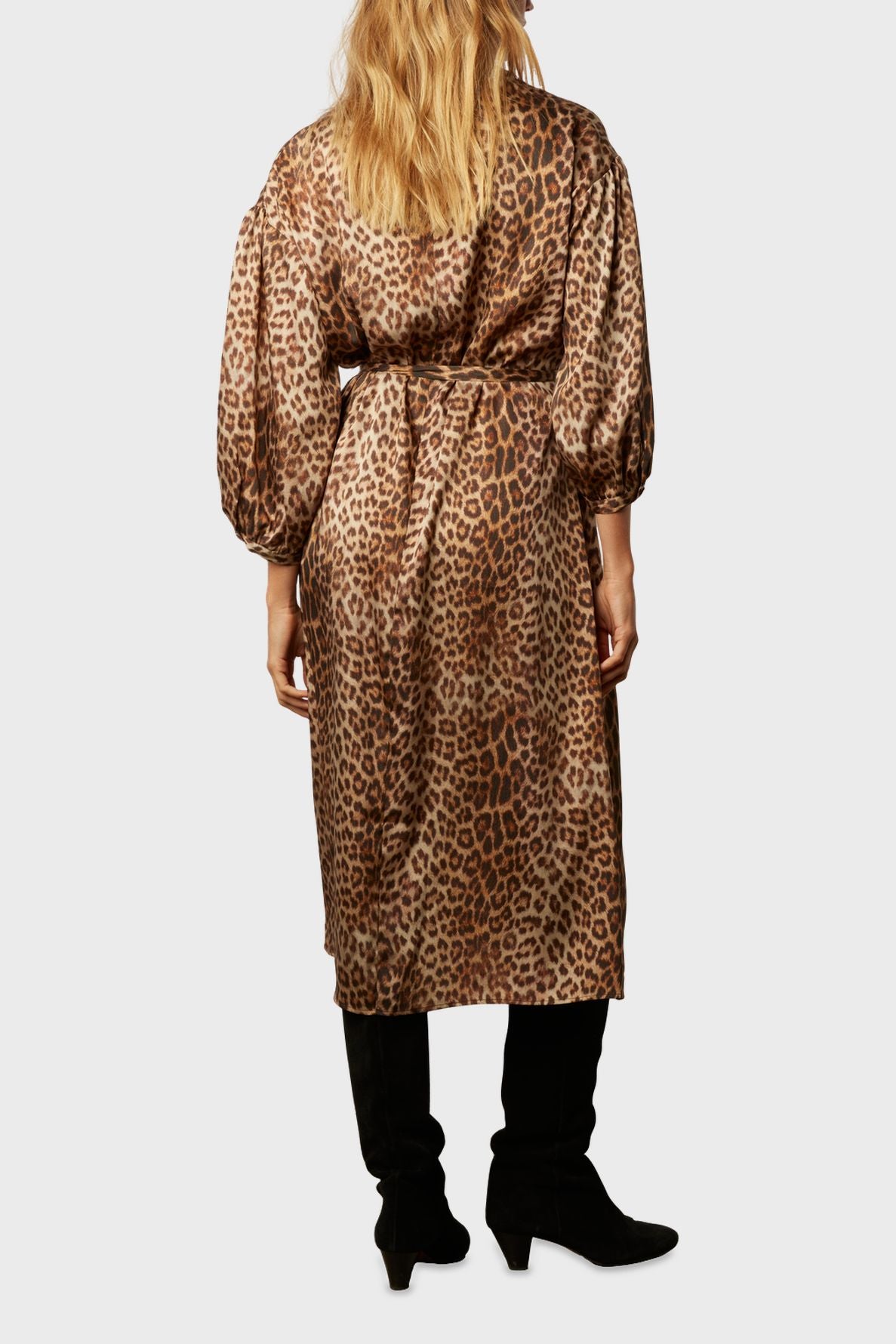 A person with long blonde hair wears a robe longue léopard, featuring long sleeves and a belted waist in a matière douce toucher soie, paired with black boots, standing with their back to the camera against a plain background.