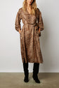 A woman wearing a robe longue léopard with long sleeves and a belt, styled with black knee-high boots, stands against a plain white background. The matière douce toucher soie of the dress adds an elegant touch to her look as she casually places her hands in her pockets, her face partially out of frame.