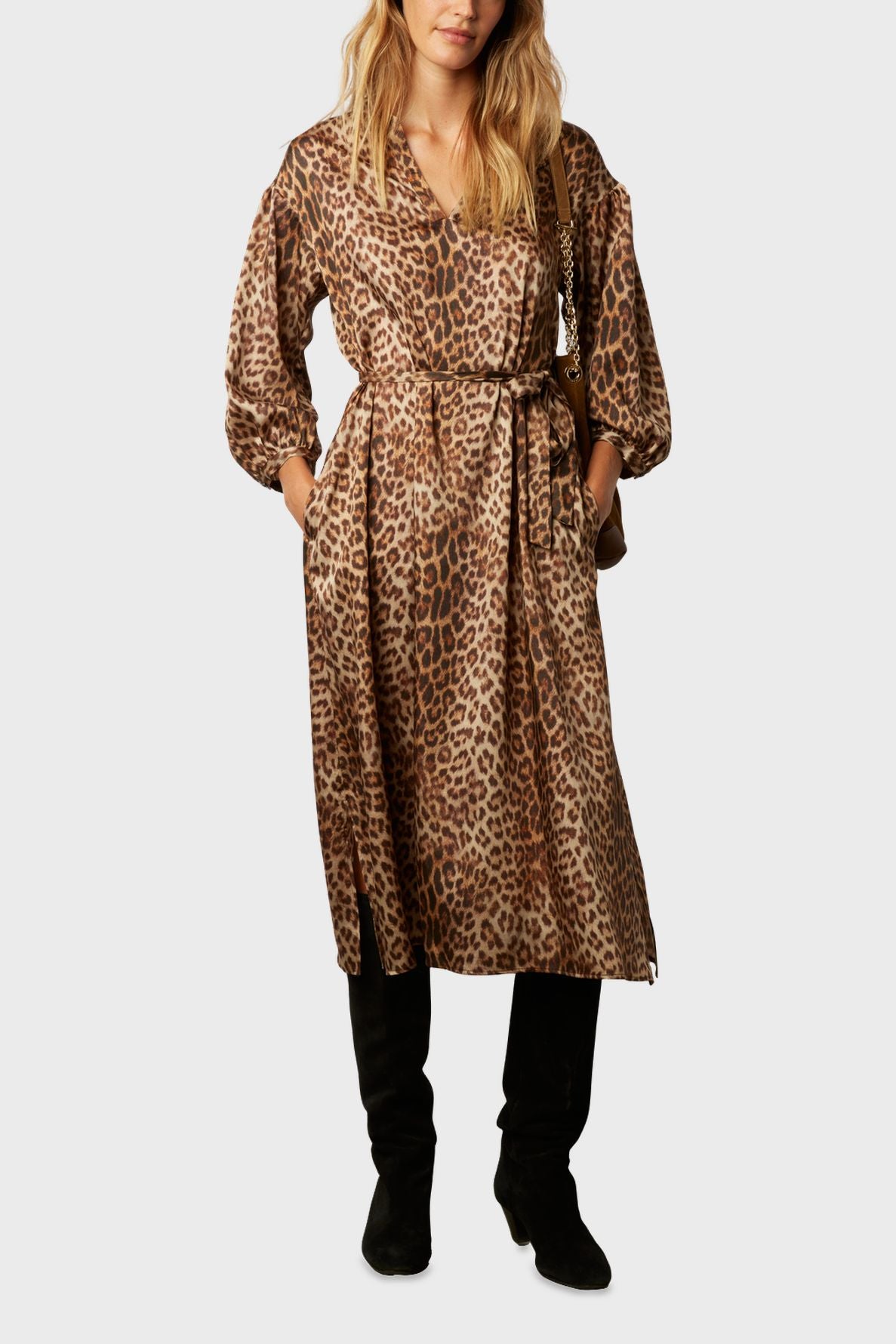A woman wearing a robe longue léopard, featuring long sleeves and a belted waist in a matière douce toucher soie, styled with black pants and black boots, stands with her hands in her pockets against a plain background.