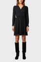 A woman wearing a long-sleeve, knee-length black dress with a V-neckline and black knee-high boots, standing against a plain light background.