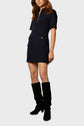 A woman wearing a short-sleeved navy dress with front pockets and a zip collar, paired with knee-high black boots, standing against a plain light background.