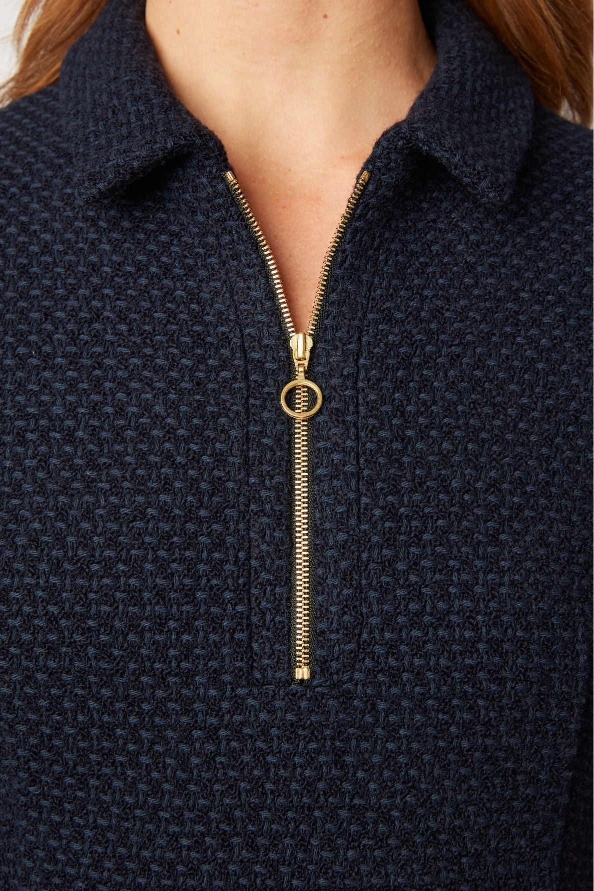 Close-up of a person wearing a navy textured zip-up sweater with a gold zipper and ring pull. Only the lower part of the face, neck, collar, and sweater are visible.