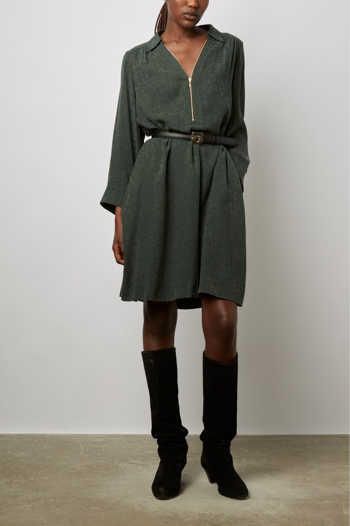 A person wearing a dark green zip-up dress with long sleeves, a black belt, and black knee-high boots stands against a plain light background.