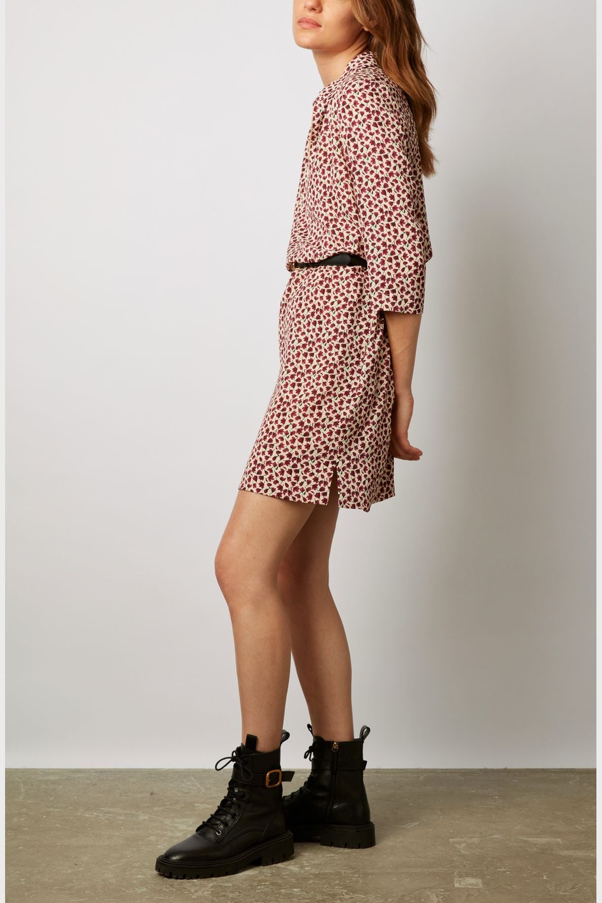 A person stands sideways wearing a pink and red patterned dress with a black belt and black lace-up boots. The background is plain and light-colored.