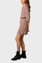 Woman in a short-sleeve, knee-length, red and white patterned dress with a black belt, standing sideways. She wears black combat boots and has long, wavy brown hair. Background is plain light gray.