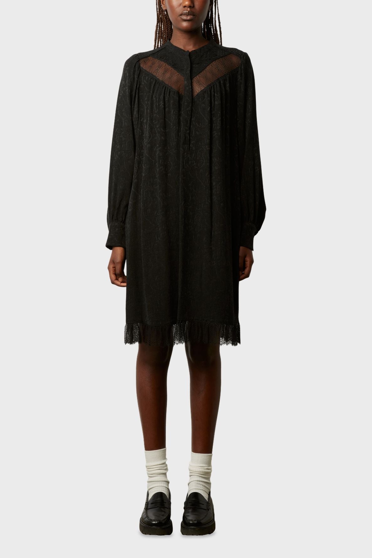 A person wearing a loose black long-sleeve dress with sheer panels at the chest and hem, reminiscent of the dentelle bohème chic style, paired with white socks and black loafers, standing against a plain light background.