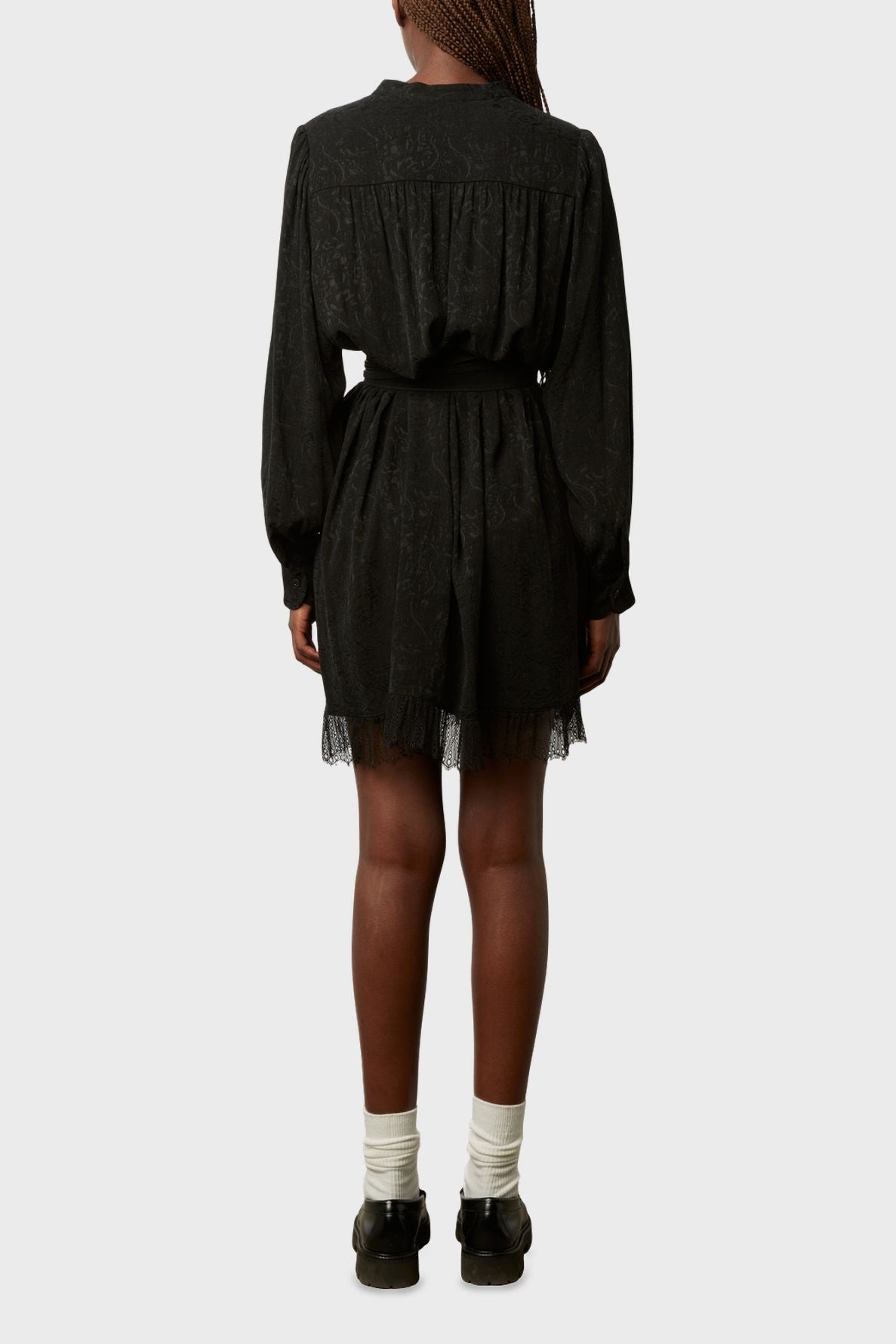 A person stands facing away, wearing a black long-sleeve dress with lace trim inspired by la dentelle bohème chic, paired with black shoes and white socks. The background is plain and light gray.