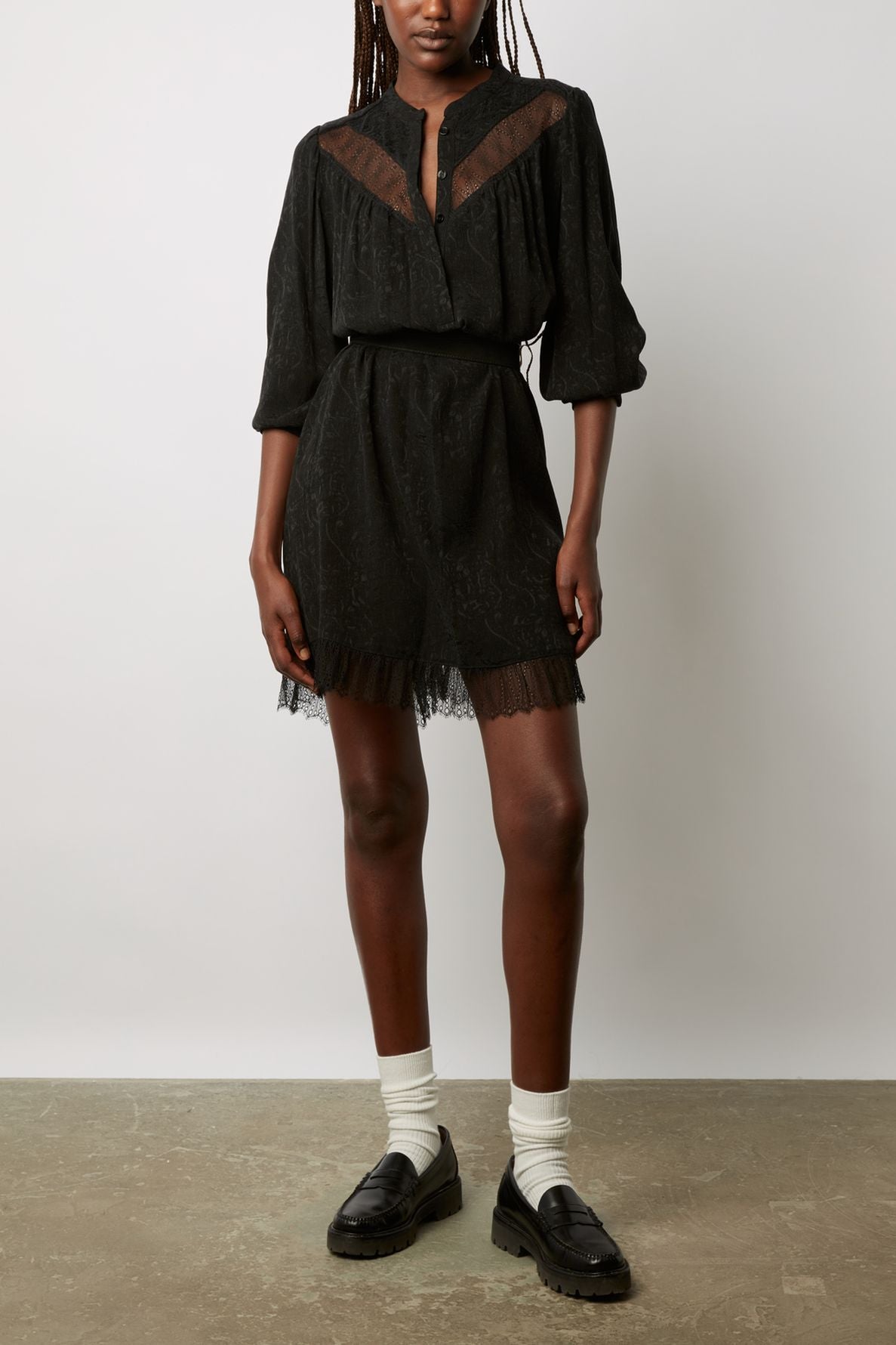 A person wearing a black long-sleeve blouse with mesh details, matching black shorts with dentelle bohème chic trim, white socks, and black loafers stands against a plain background.