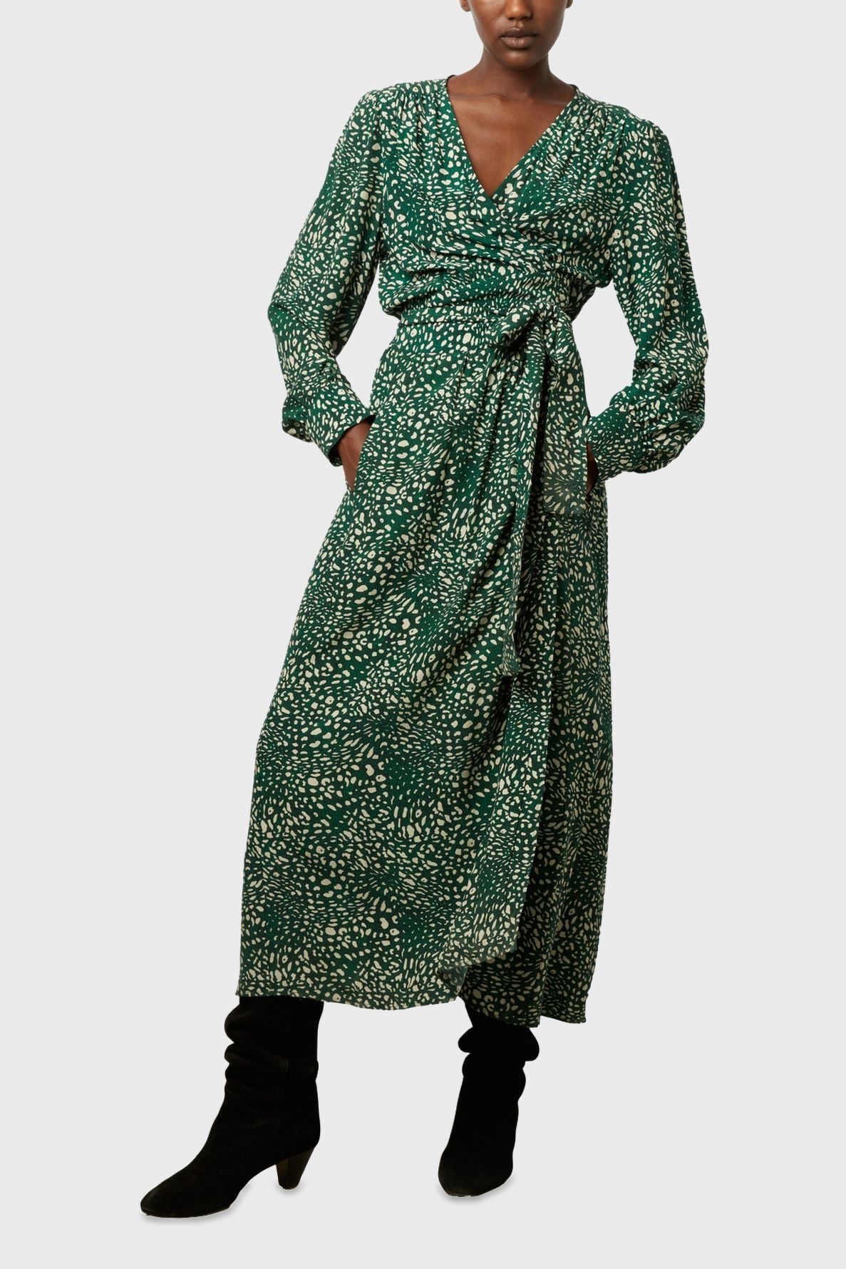 Person wearing a long-sleeved, green wrap dress with a white dotted pattern and a tie at the waist, paired with black heeled boots, standing against a plain light background.