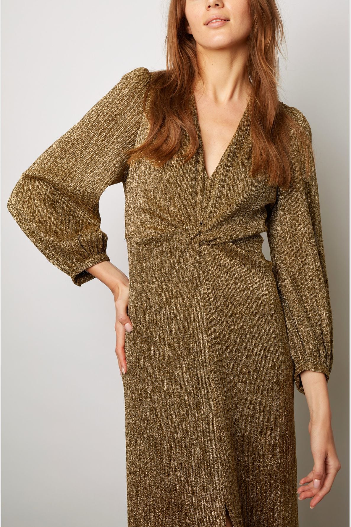 Woman in a long-sleeve, gold shimmering robe longue with a V-neckline, standing against a plain light background, hand on her hip. Her face is partially out of view—an elegant pose in this radiant robe de soirée en lurex doré.