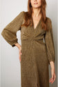 Woman in a long-sleeve, gold shimmering robe longue with a V-neckline, standing against a plain light background, hand on her hip. Her face is partially out of view—an elegant pose in this radiant robe de soirée en lurex doré.