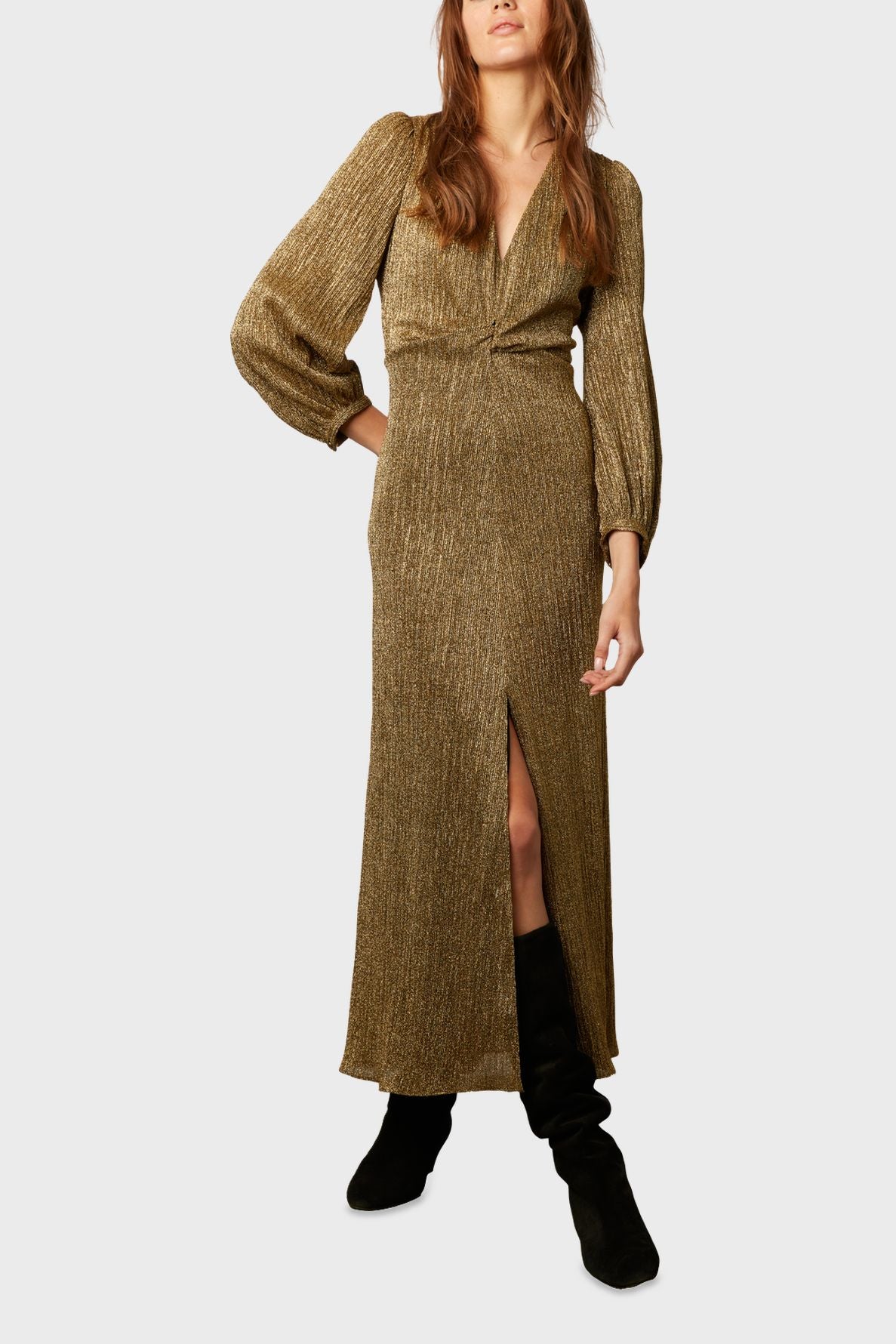 Woman wearing a long, gold, shimmery lurex doré robe de soirée with long sleeves and a front slit, paired with black knee-high boots. Her face is partially visible, and she stands against a plain light background.