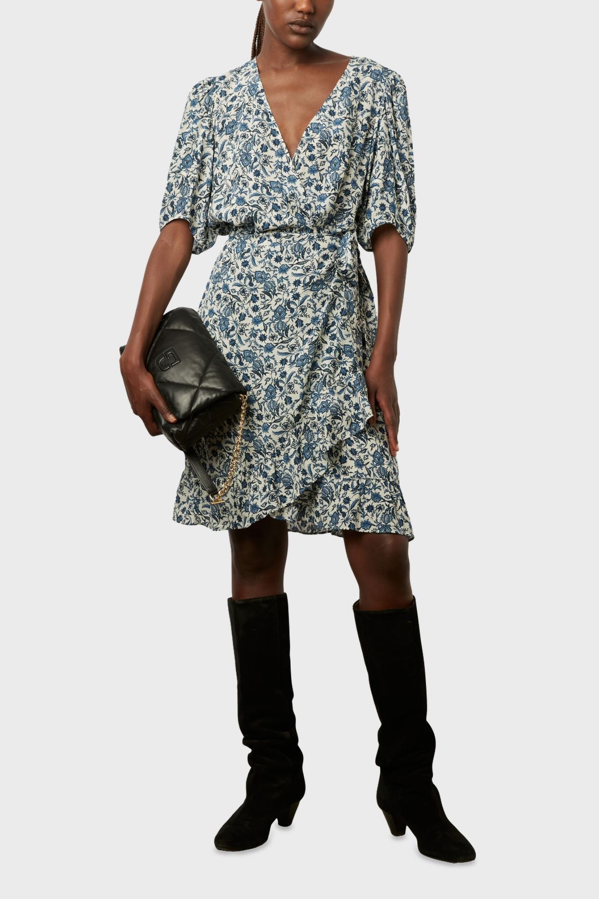 A person wearing a blue and white floral wrap dress, black knee-high boots, and holding a black quilted handbag with a chain strap, standing against a plain background.