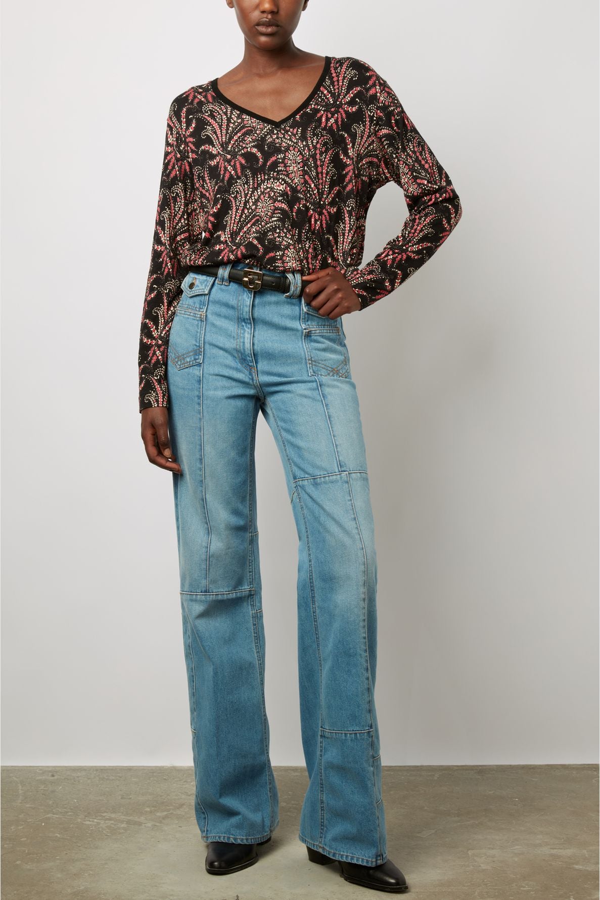Person wearing a black long-sleeve shirt with a pink and white leafy pattern, tucked into high-waisted light blue jeans with visible seams, black belt, and black shoes, standing against a plain background.