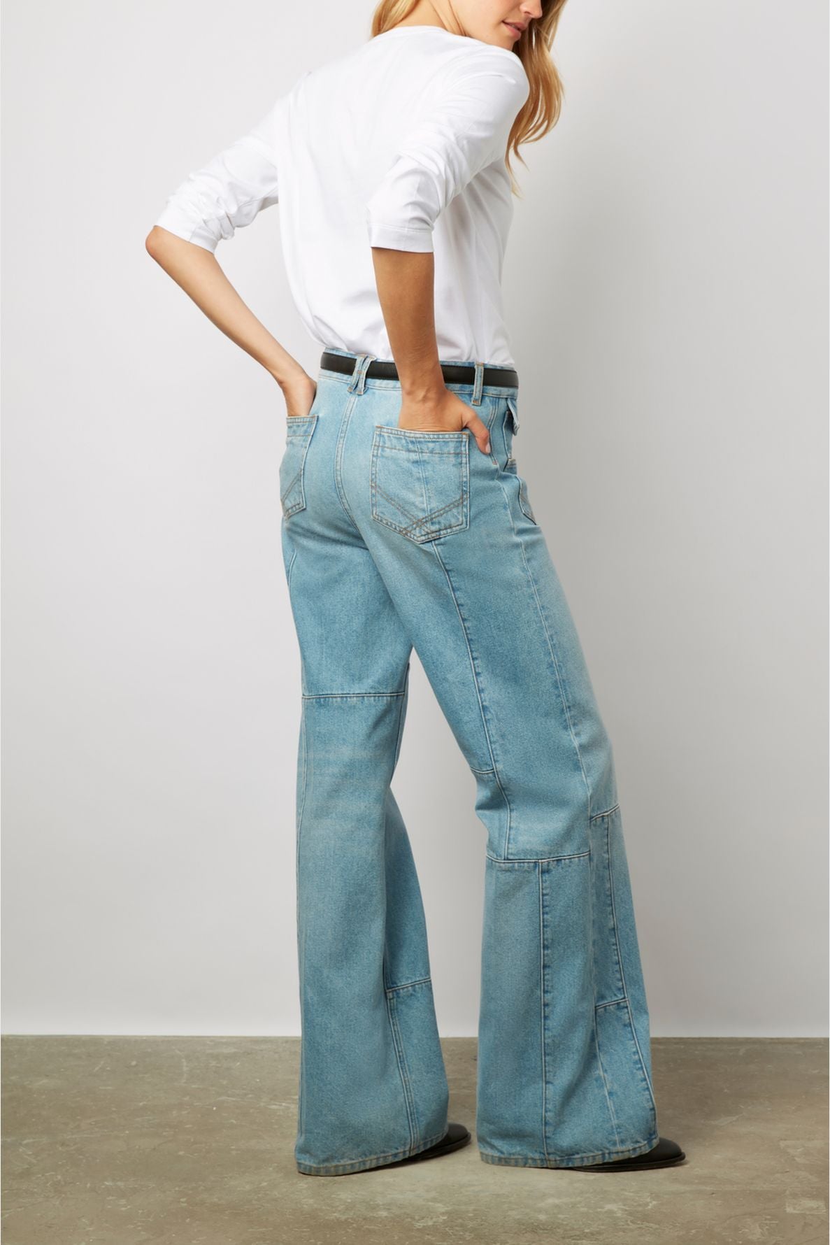 A person stands against a plain background wearing a white long-sleeve shirt, light blue high-waisted wide-leg jeans, and a black belt. They have one hand in a back pocket and are turned slightly to the side.