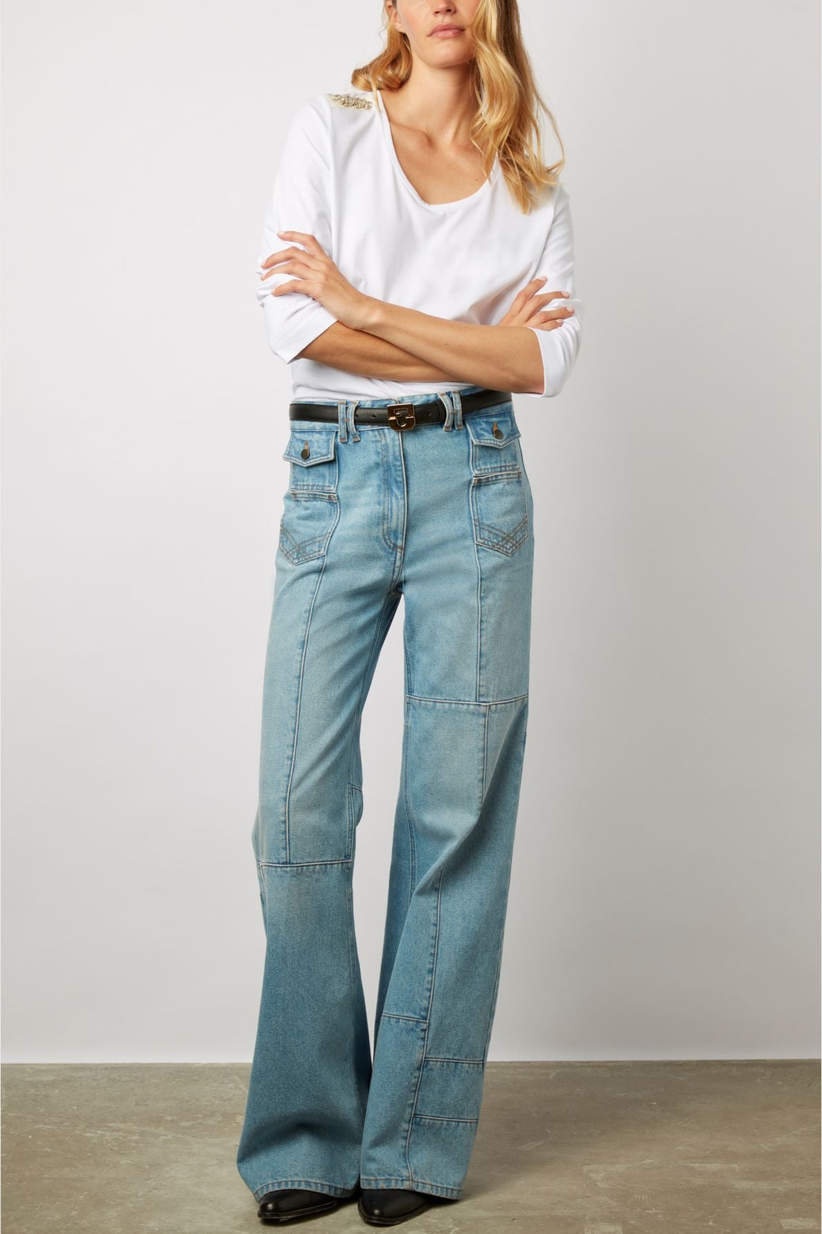 A woman stands with arms crossed, wearing a white long-sleeve top and high-waisted, wide-leg blue jeans with front pockets and patches, paired with black shoes.