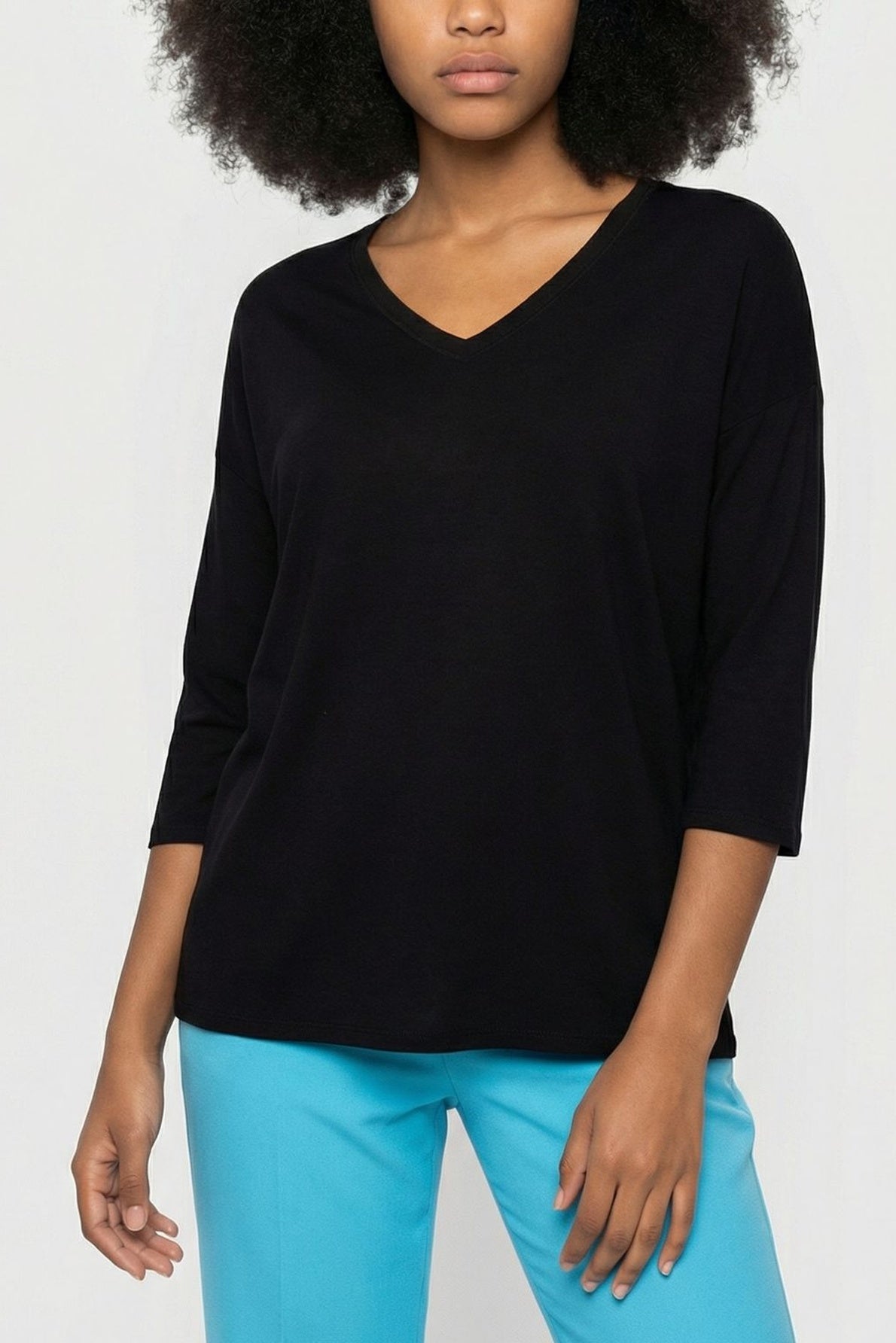 A woman wearing a loose black V-neck top with three-quarter sleeves and bright blue pants stands against a plain light background.