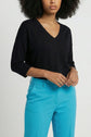 A person wearing a black V-neck top with three-quarter sleeves and bright blue pants, standing against a plain light background. Their hands are clasped at the waist.