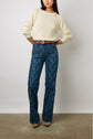 A person stands against a white wall, wearing a cream sweater, high-waisted blue patterned jeans, a brown belt, and beige pointed shoes. Their arms are bent, and hands rest on their hips.