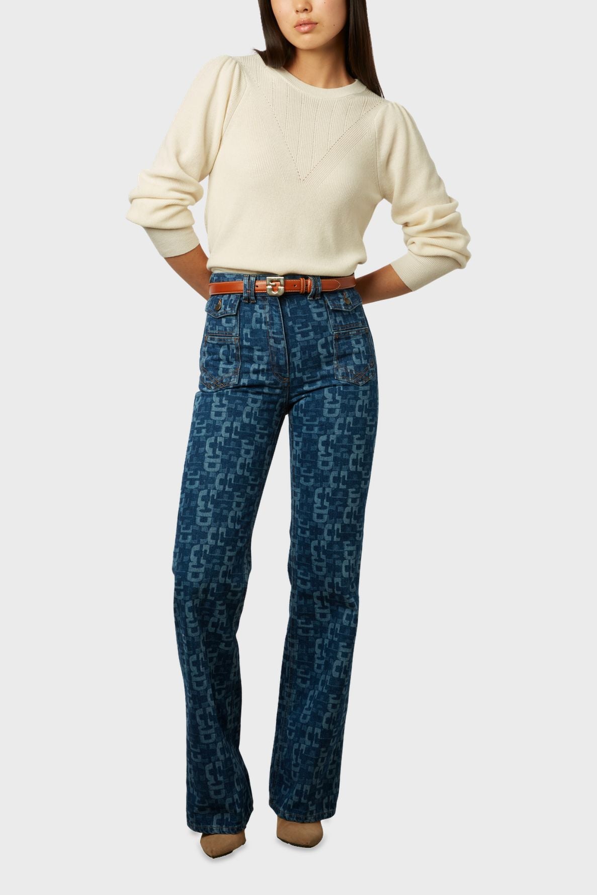 Woman wearing a cream sweater, patterned high-waisted blue jeans with front pockets, a brown belt, and brown shoes, posing with hands on hips against a plain light background.