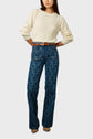 Woman wearing a cream sweater, patterned high-waisted blue jeans with front pockets, a brown belt, and brown shoes, posing with hands on hips against a plain light background.