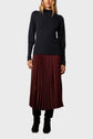 Woman wearing a long-sleeve black sweater with a burgundy pleated midi skirt and black boots, standing against a plain light background.