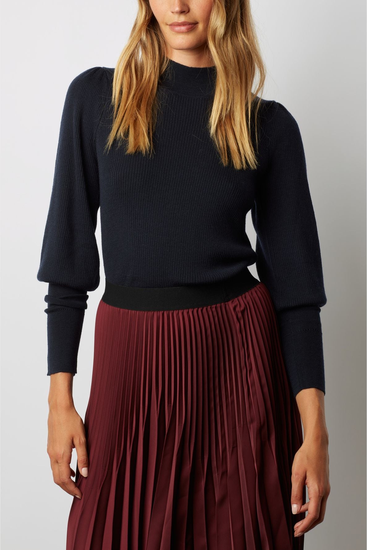 Woman wearing a long-sleeve, dark navy sweater and a high-waisted, pleated burgundy skirt, standing against a plain light background. Her hair is loose and wavy, and her face is partially out of frame.