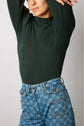A person wearing a dark green ribbed turtleneck and high-waisted blue jeans with a pattern poses with arms raised, partially covering their face.