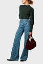 A woman wearing a dark green long-sleeve top and high-waisted, patterned blue jeans stands holding a burgundy handbag. She is looking to the side, and her hair is tied back.