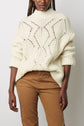 A person wearing a cream-colored, chunky knit pull torsades with a geometric pattern and tan pants, standing against a plain light background.