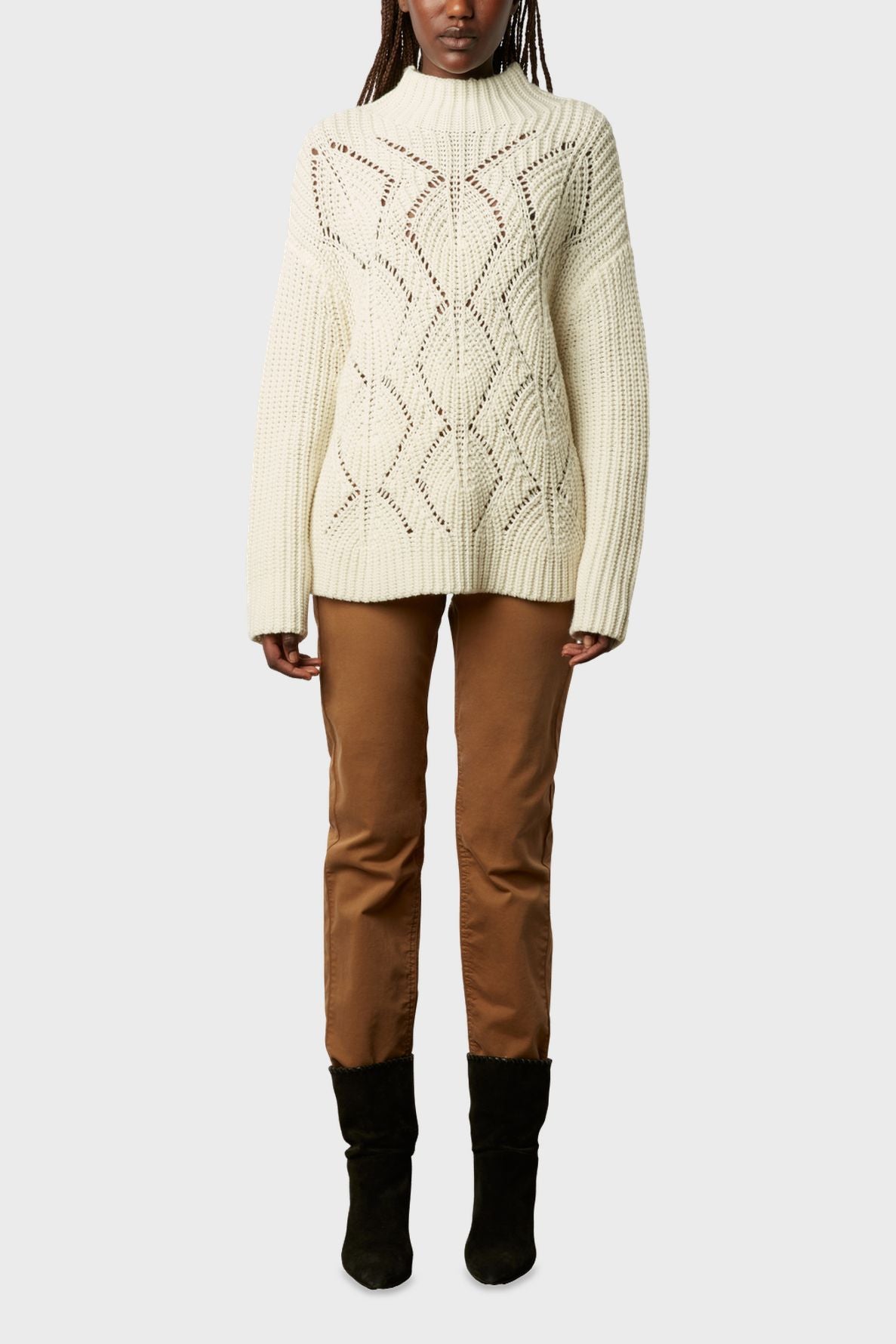 A person is wearing a cream-colored pull col montant en laine mélangée with a diamond pattern, brown pants, and black boots, standing against a plain light gray background.