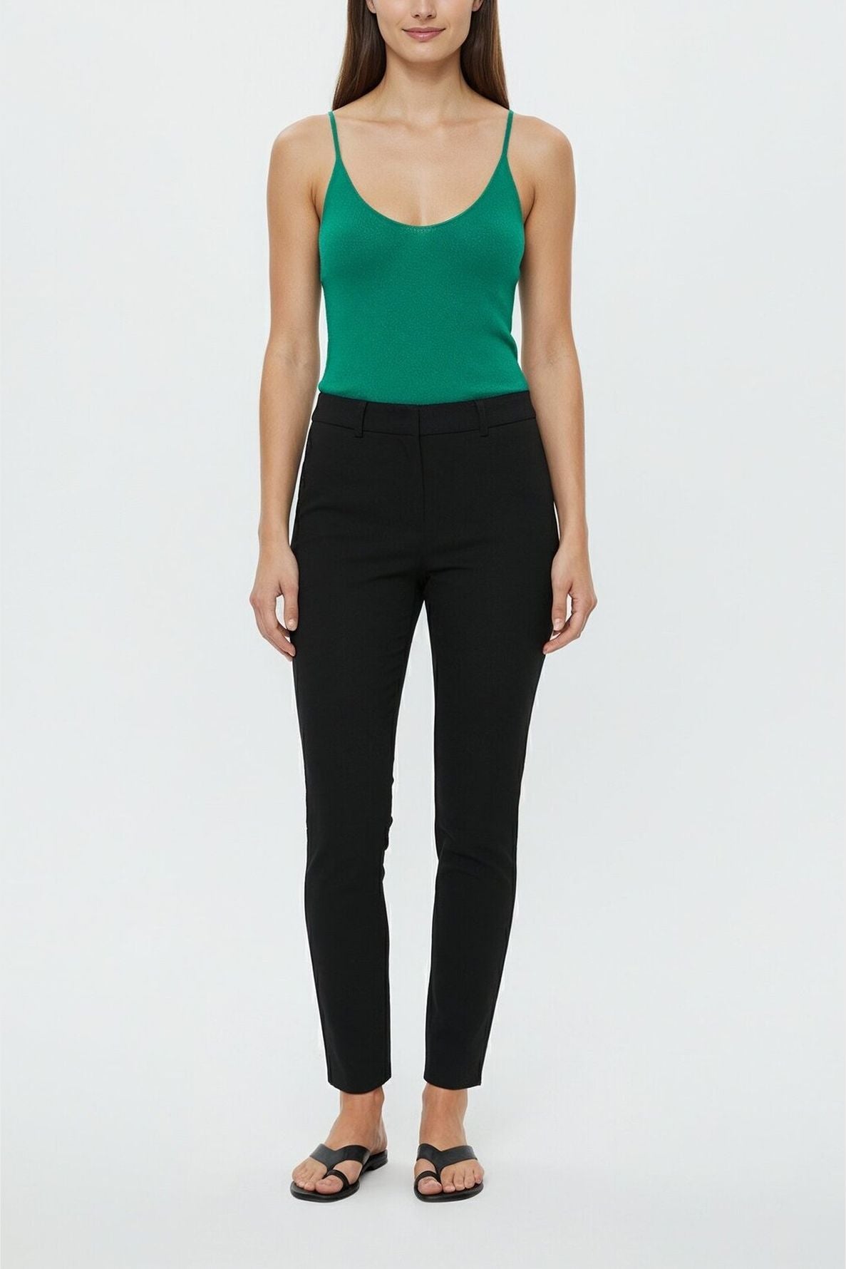 A person stands facing forward, wearing a green débardeur femme paired with black fitted pants and black sandals, against a plain light background.