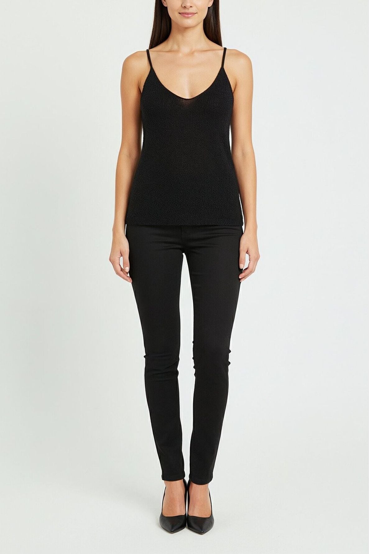 A woman stands facing forward, wearing a sleek débardeur noir paired with black skinny pants and black pointed-toe heels against a plain white background.