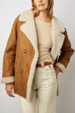 Woman wearing a tan manteau court en peau lainée mouton with large cream lapels, a beige sweater, and high-waisted cream pants. She stands against a plain background, with one hand in her pocket.