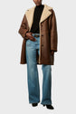 Person wearing a brown manteau peau de mouton with a cream collar, black turtleneck sweater, blue wide-leg jeans, and black shoes, standing against a light background.