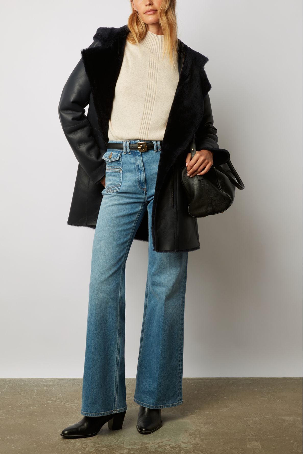Woman wearing a cream sweater, flared blue jeans, black heeled boots, and a black manteau en peau lainée with a furry collar. She holds a black handbag at her side. The background is plain and neutral.