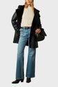 A person wearing a cream sweater, a black manteau en peau lainée with a furry collar, blue wide-leg jeans, black boots, and holding a black handbag.