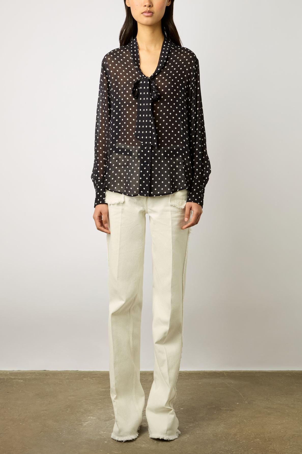 A person wearing a sheer black blouse with white polka dots and a bow at the neck, paired with high-waisted, off-white straight-leg pants with frayed hems, standing against a plain background.
