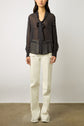 A person wearing a sheer black blouse with white polka dots and a bow at the neck, paired with high-waisted, off-white straight-leg pants with frayed hems, standing against a plain background.