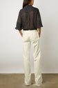 A person stands facing away, wearing a sheer black blouse with white polka dots and white high-waisted, wide-legged jeans with frayed hems. The background is a plain white wall and concrete floor.