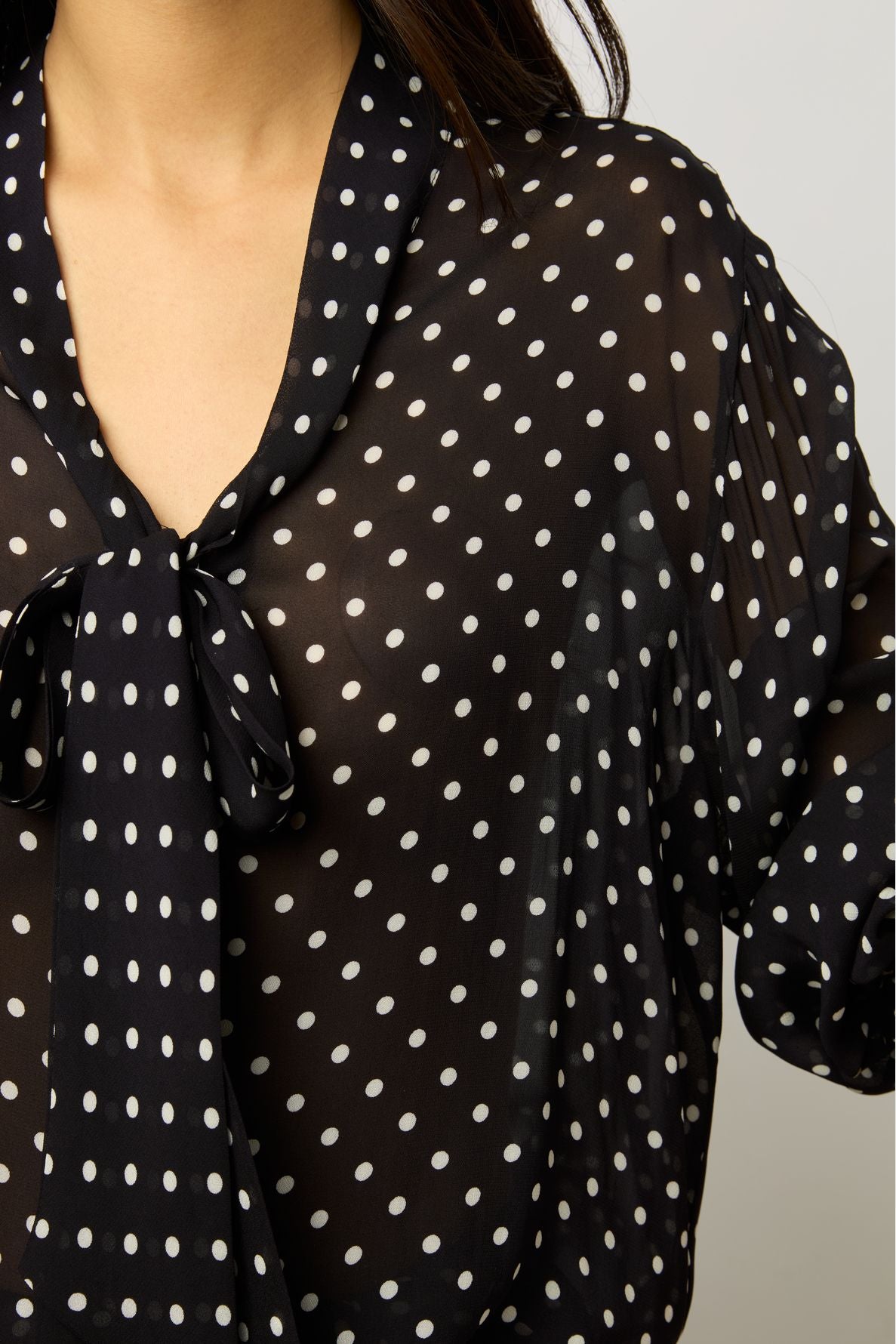 A person wearing a sheer black blouse with white polka dots and a tied bow at the collar. Only the upper torso and part of one arm are visible.